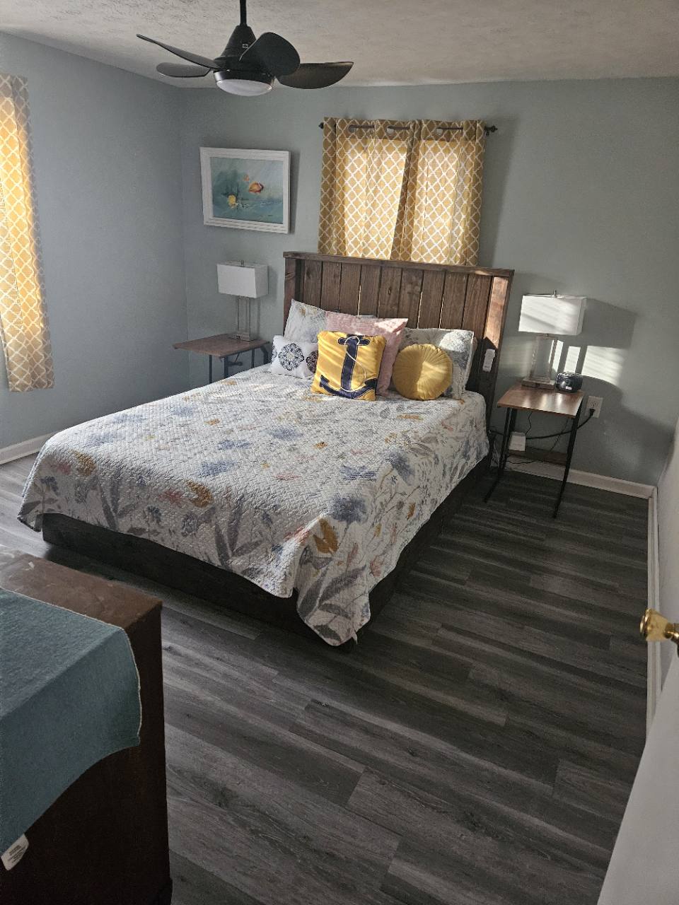 Primary bedroom queen bed with patterned bedding, anchor cushion, wooden headboard and bedside lamps